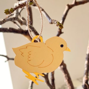 Op de afbeelding: Een geel kuiken ornament met een lus van touw om op te hangen. Het kuiken is gemaakt van metaal en heeft een eenvoudig, cartoonachtig ontwerp.