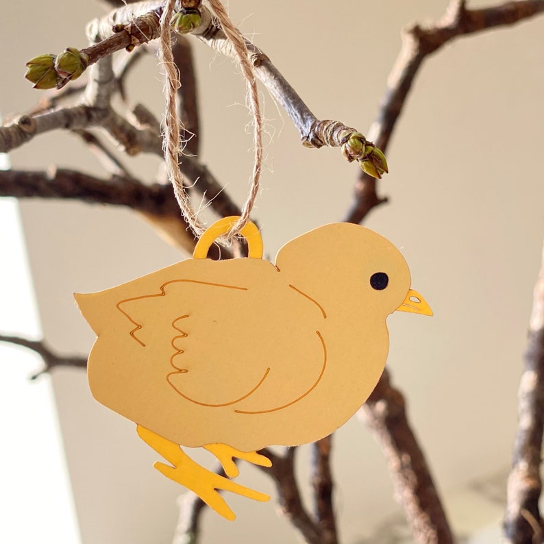 Op de afbeelding: Een geel kuiken ornament met een lus van touw om op te hangen. Het kuiken is gemaakt van metaal en heeft een eenvoudig, cartoonachtig ontwerp.