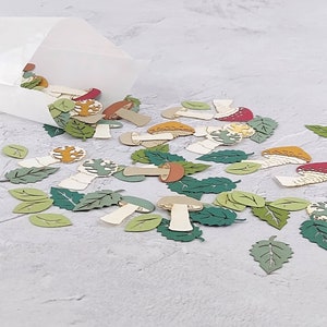 May include: A scattering of paper cutouts of mushrooms and leaves in various shades of green, brown, red, and yellow. The cutouts are scattered on a white surface.
