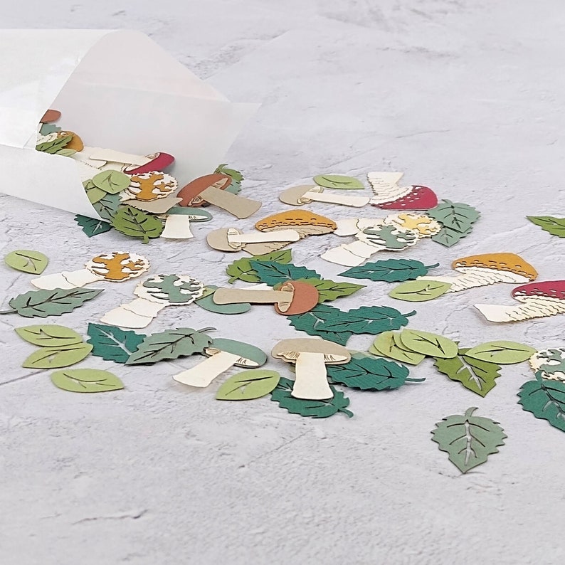 May include: A scattering of paper cutouts of mushrooms and leaves in various shades of green, brown, red, and yellow. The cutouts are scattered on a white surface.