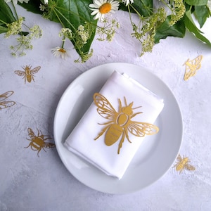 May include: A white napkin with a gold bee napkin ring on a white plate. The plate is surrounded by small gold bee cutouts on a white background.