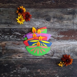 May include: A stack of colorful paper diya lamps with yellow and orange flames, arranged on a rustic wooden surface. The lamps are in shades of green, red, blue, and purple. Decorative flowers are scattered around the lamps.