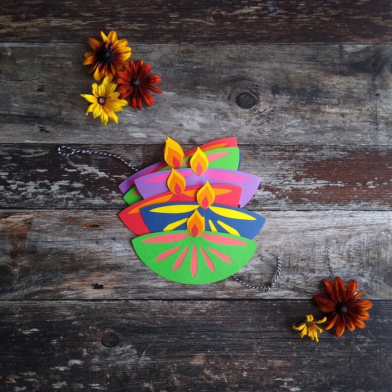 May include: A stack of colorful paper diya lamps with yellow and orange flames, arranged on a rustic wooden surface. The lamps are in shades of green, red, blue, and purple. Decorative flowers are scattered around the lamps.
