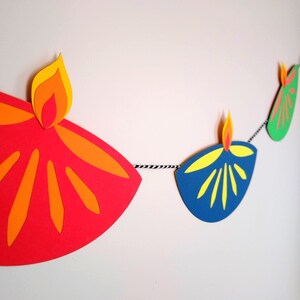 May include: A colorful Diwali banner featuring paper cutouts of oil lamps in red, orange, yellow, blue, and green. The lamps are strung together with a black and white twisted cord, creating a festive decoration.