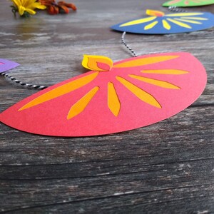 May include: A colorful paper garland featuring a series of stylized flame shapes in red, orange, yellow, blue, green, and purple. The garland is strung on a black and white twine, perfect for festive decorations.