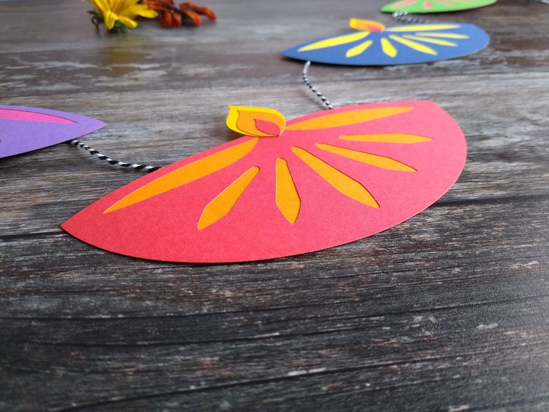 May include: A colorful paper garland featuring a series of stylized flame shapes in red, orange, yellow, blue, green, and purple. The garland is strung on a black and white twine, perfect for festive decorations.