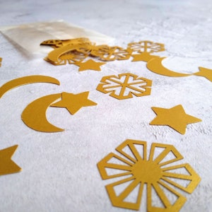 May include: Gold paper cutouts of stars, moons, and snowflakes spill from a small paper bag. The shapes are scattered on a light gray surface, creating a festive and decorative arrangement. Ideal for crafting or party decorations.