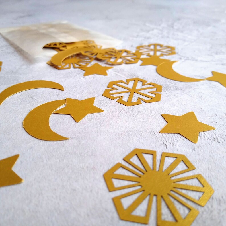 May include: Gold paper cutouts of stars, moons, and snowflakes spill from a small paper bag. The shapes are scattered on a light gray surface, creating a festive and decorative arrangement. Ideal for crafting or party decorations.