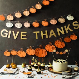 May include: A Thanksgiving-themed banner with the words "GIVE THANKS" in gold letters, decorated with orange and brown paper pumpkins. A white frosted cake with black and white sprinkles sits on a wooden board in front of the banner. The cake is surrounded by miniature cupcakes with various toppings.