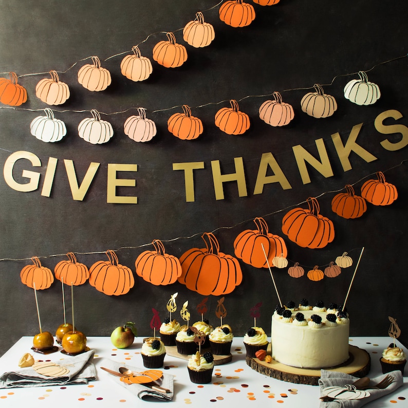 May include: A Thanksgiving-themed banner with the words "GIVE THANKS" in gold letters, decorated with orange and brown paper pumpkins. A white frosted cake with black and white sprinkles sits on a wooden board in front of the banner. The cake is surrounded by miniature cupcakes with various toppings.