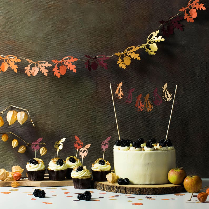 May include: A white frosted cake decorated with blackberries and topped with a fall-themed paper garland. The cake is surrounded by six mini cupcakes with fall-themed paper flags. The cupcakes are on a wooden surface with a brown background.