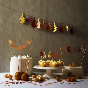 May include: A white cake with fall-themed decorations, including a garland of paper pumpkins and a string of paper leaves. The cake is surrounded by cupcakes with fall-themed decorations, including paper leaves and paper pears. The cupcakes are on a white cake stand. The background is a gray wall with a garland of paper leaves.