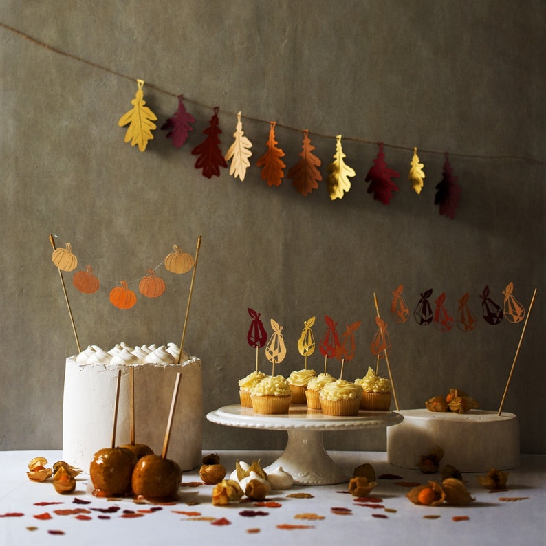 May include: A white cake with fall-themed decorations, including a garland of paper pumpkins and a string of paper leaves. The cake is surrounded by cupcakes with fall-themed decorations, including paper leaves and paper pears. The cupcakes are on a white cake stand. The background is a gray wall with a garland of paper leaves.