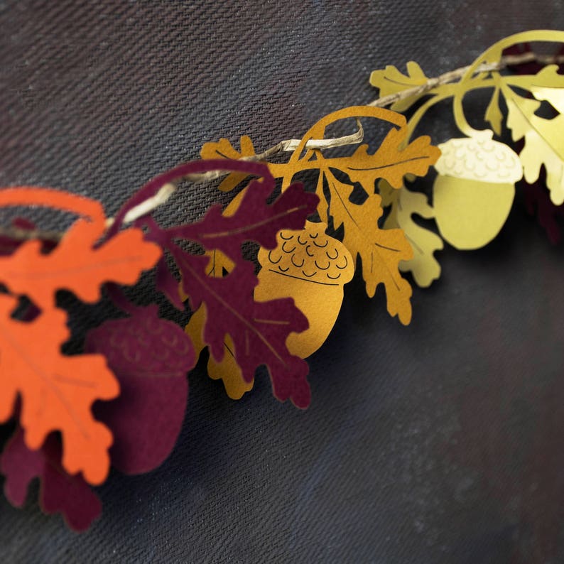 May include: A paper garland with orange, burgundy, and yellow leaves and acorns. The garland is strung with twine and is perfect for fall decor.