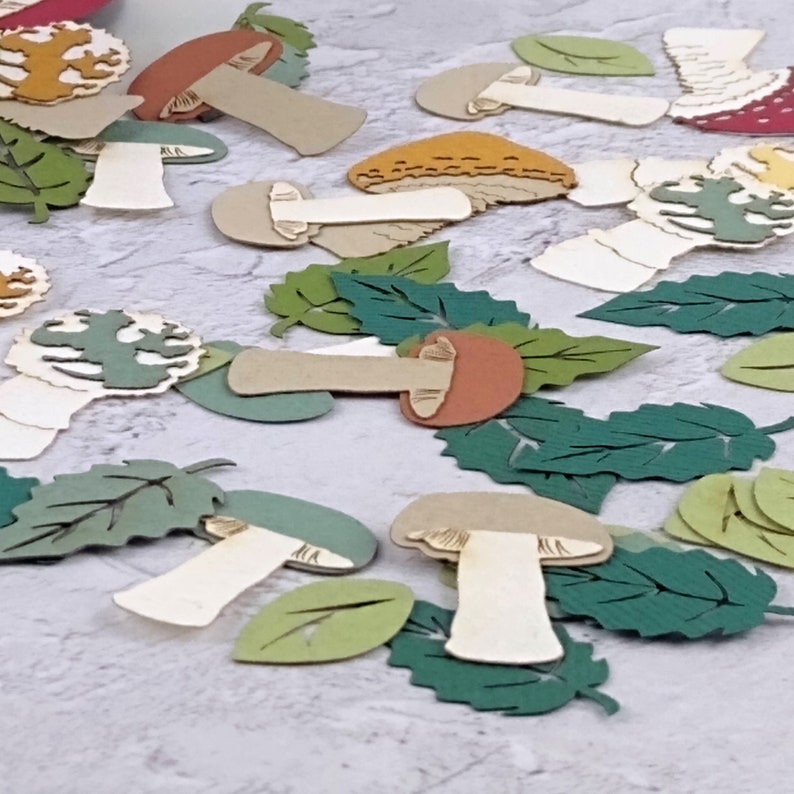 May include: Assortment of paper craft mushrooms and leaves in various shades of green, brown, and white. The mushrooms have different shapes and sizes, and the leaves are cut in various shapes.