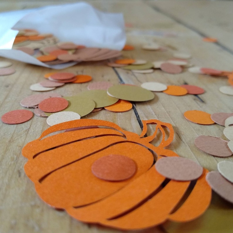 May include: Orange paper pumpkin cutout with brown and orange paper circles scattered around it.