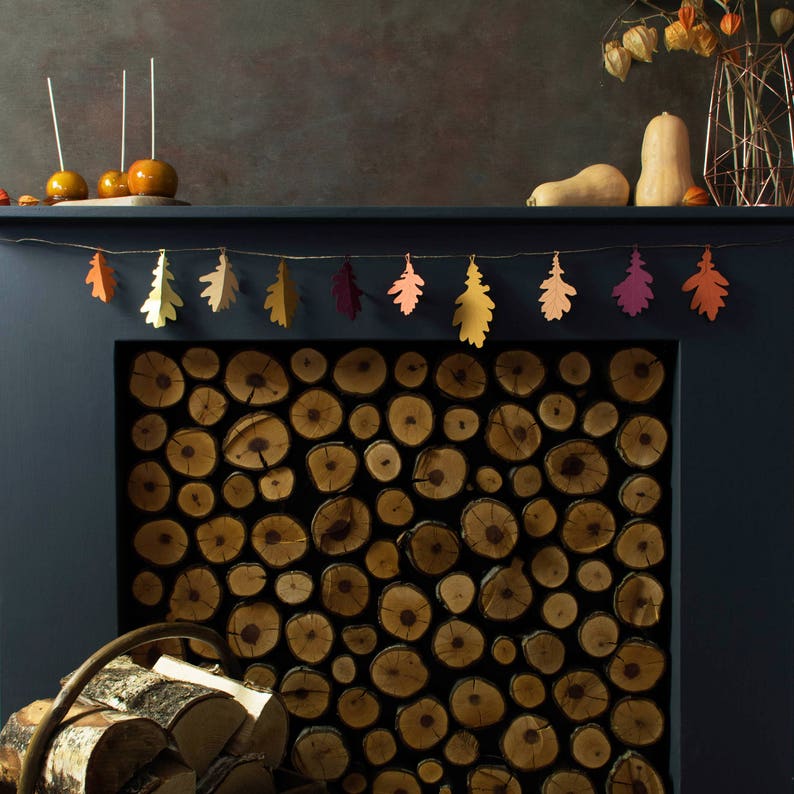May include: A dark blue fireplace mantel with a garland of paper leaves in shades of orange, yellow, and red. The fireplace is filled with stacked logs of wood.