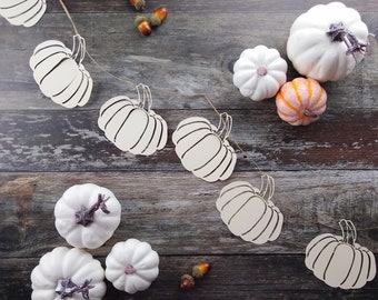 May include: An assortment of fall-themed decorations, including white and orange pumpkins of various sizes, along with decorative pumpkin cutouts and acorns. The pumpkins have textured surfaces and are arranged on a rustic wooden background.