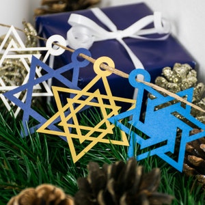 Closeup of four star of david paper decorations. Each of the white, navy blue, gold and electric blue star shapes have different geometric shapes cut out to make 4 intricate star shapes