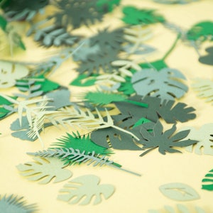 May include: A collection of green and blue paper leaves scattered on a yellow background. The leaves are cut in various shapes and sizes, resembling tropical foliage.