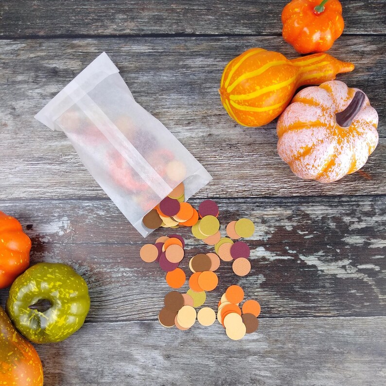May include: A white paper bag filled with colorful confetti circles in shades of orange, yellow, brown, and red. The bag is spilling out onto a wooden surface with three gourds in the background.