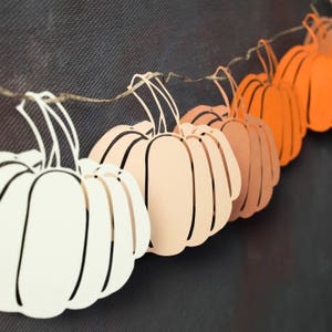 May include: A string of paper pumpkins in various shades of orange, peach, and white. The pumpkins are cut out and strung together with twine.