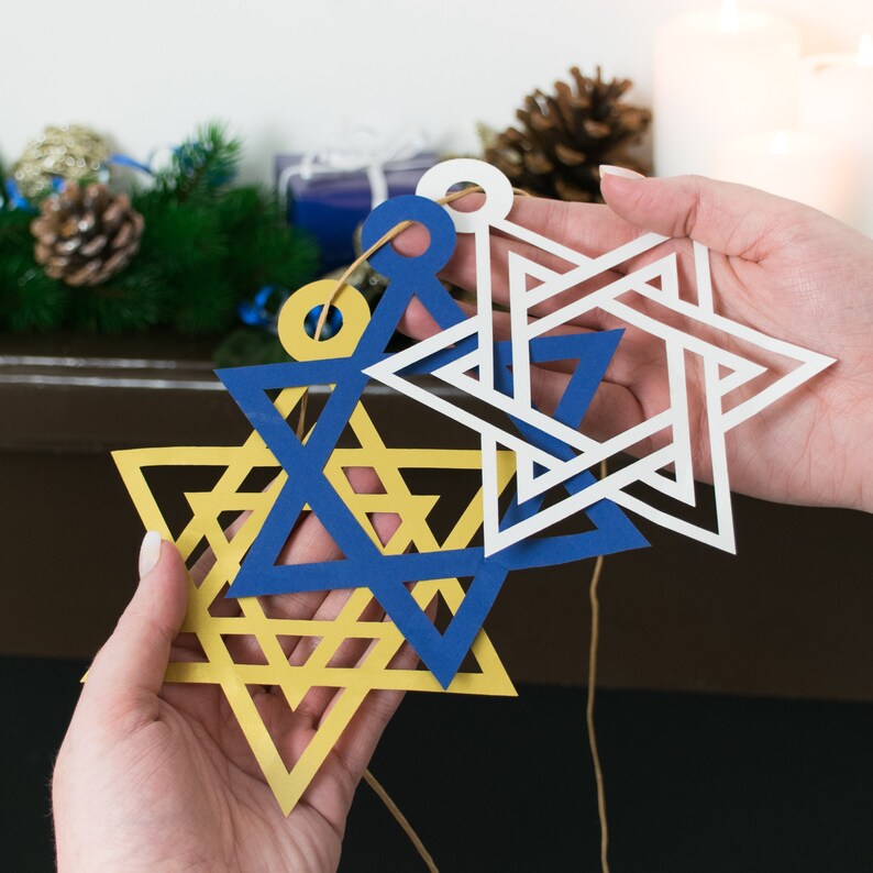 closeup of 3 large star of david decorations held in models hands. each star is approximately the size of model's palm. Stars are cut in gold, white and navy blue papers.