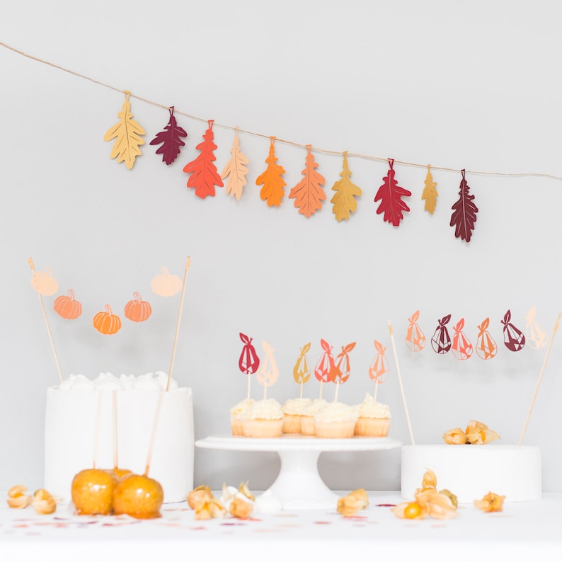 May include: A fall-themed party table setting with cupcakes topped with pear-shaped decorations, caramel apples, and a white cake stand. A garland of orange, red, and yellow paper leaves hangs above the table.