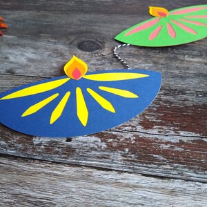 May include: A festive garland featuring paper cutouts of oil lamps. The lamps are in blue and green, with yellow and pink accents. The garland is strung on a black and white string, perfect for Diwali celebrations.