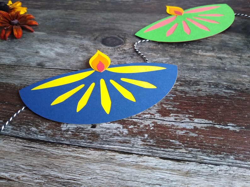 May include: A festive garland featuring paper cutouts of oil lamps. The lamps are in blue and green, with yellow and pink accents. The garland is strung on a black and white string, perfect for Diwali celebrations.