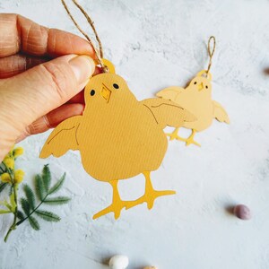 Op de afbeelding: Twee gele papieren kuikenversieringen met touwtjes om op te hangen. De kuikens hebben zwarte ogen en snavels. De kuikens zijn op een witte achtergrond met gele bloemen en gevlekte eieren.