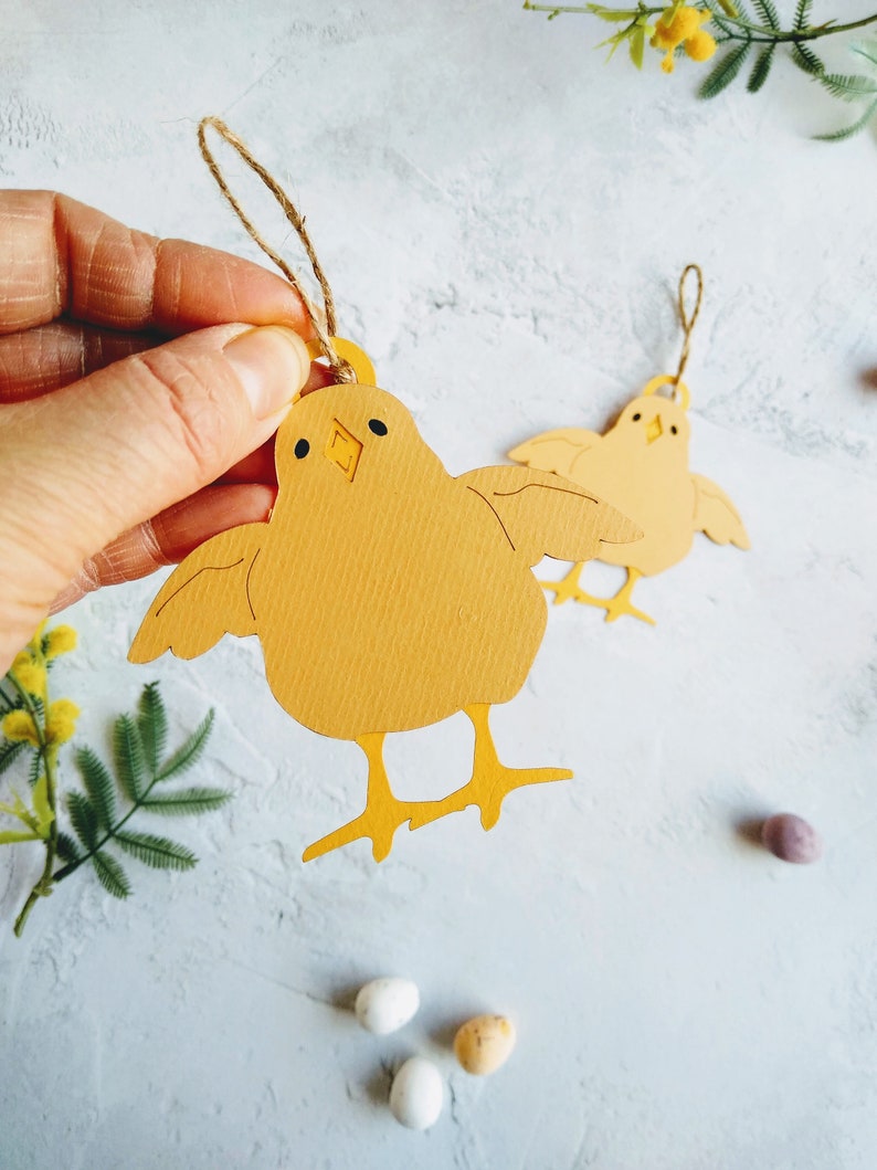 Op de afbeelding: Twee gele papieren kuikenversieringen met touwtjes om op te hangen. De kuikens hebben zwarte ogen en snavels. De kuikens zijn op een witte achtergrond met gele bloemen en gevlekte eieren.