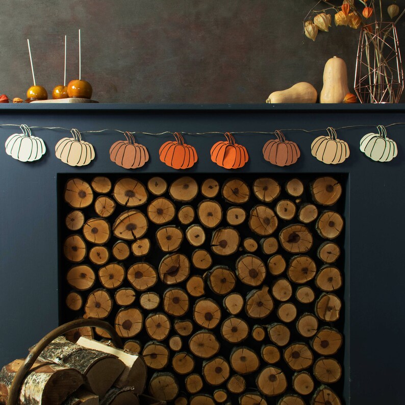 May include: A fireplace mantel decorated with a garland of paper pumpkins in shades of orange, peach, and cream. The fireplace is stacked with logs and has a dark blue paint finish.