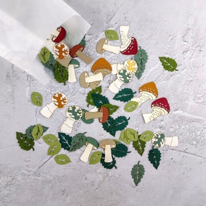 May include: A white paper bag spills out a variety of colorful paper mushroom cutouts. The mushrooms are in various shapes and sizes, with red, orange, yellow, and brown accents. The cutouts are scattered on a light gray background.