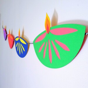 May include: A colorful paper garland featuring stylized oil lamps. Each lamp is a different color, including green, blue, red, and purple, with pink and yellow flame details. The garland is strung on a black and white string, perfect for festive decorations.