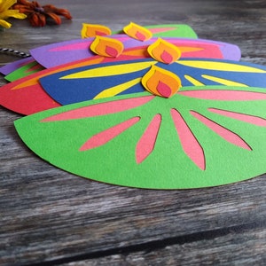 May include: Colorful paper cutouts in the shape of oil lamps, with flames on top. The lamps are in shades of green, red, purple, and blue, with yellow and pink accents. A black and white string is attached.
