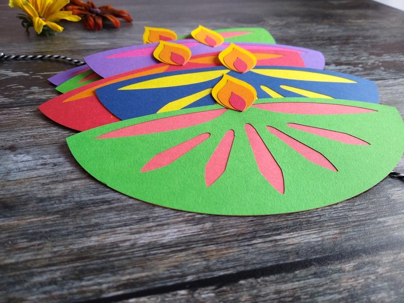 May include: Colorful paper cutouts in the shape of oil lamps, with flames on top. The lamps are in shades of green, red, purple, and blue, with yellow and pink accents. A black and white string is attached.
