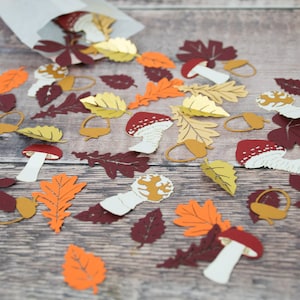 May include: A scattering of paper cutouts of autumn leaves and mushrooms in shades of red, orange, yellow, and brown. The cutouts are on a woodgrain background.