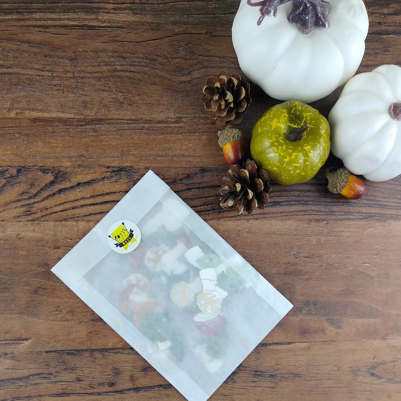 May include: A white envelope with a yellow sticker that says "Good Vibes Only" on it. The envelope is on a wooden surface with a green apple, white gourds, and pine cones.