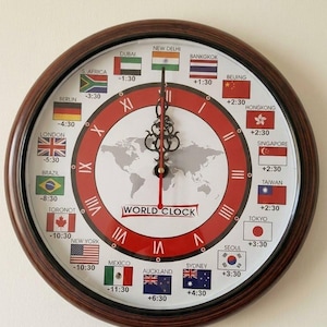 Puede incluir: Reloj mundial redondo con marco de madera marrón oscuro. La esfera del reloj presenta un diseño rojo y blanco con un mapa del mundo y banderas que representan diferentes zonas horarias. Las manecillas del reloj son negras.