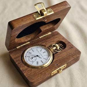 Op de afbeelding: Een goudkleurig zakhorloge met een witte wijzerplaat, zwarte Romeinse cijfers en een kleine sub-wijzerplaat, rustend in een rechthoekige donkerbruine houten doos. De doos is voorzien van een messingkleurige metalen sluiting en scharnieren.