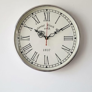 Puede incluir: Un reloj de pared redondo con marco de metal plateado. La esfera es blanquecina con números romanos y manecillas negras. Las palabras "Colonial Clock estd." y el año "1937" están impresos en la esfera.
