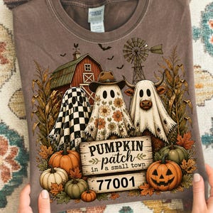 Könnte beinhalten: Ein braunes T-Shirt mit einer Grafik von drei Geistern, einer mit Karomuster, einer mit Blumenmuster und Cowboyhut und einer in Kuhform. Das Design zeigt eine Scheune, eine Windmühle, Kürbisse und den Text PUMPKIN patch in a small town 77001.
