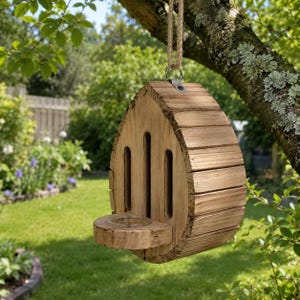 Op de afbeelding: Een druppelvormig insectenhotel van hout met een natuurlijke lichtbruine afwerking, verticale latten en een klein rond platform aan de basis. De structuur is opgehangen aan een dik touw van jute dat aan een metalen ring aan de bovenkant vastzit.