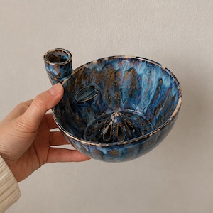 May include: A ceramic citrus juicer bowl featuring a deep blue and brown mottled glaze. The bowl includes a central reamer cone with drainage holes and a small cylindrical handle on the side, showcasing a glossy finish and varied color patterns.