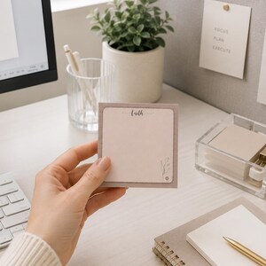 May include: A hand holding a square, light pink sticky note with the word "faith" in a handwritten font. The note has a light brown border and a small floral design in the corner. The note is on a white desk with a computer, keyboard, and other office supplies.
