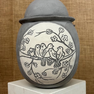 Könnte beinhalten: Eine graue Keramikurne mit abgerundetem Deckel und matter Oberfläche. Die Vorderseite zeigt ein weißes, kreisförmiges Einlegefeld mit einer detaillierten, eingeritzten Illustration von fünf Vögeln auf einem blühenden Zweig.
