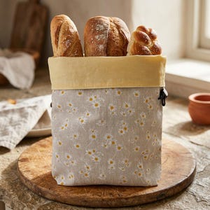 Könnte beinhalten: Ein Brotkorb aus Stoff mit hellgrauer Außenseite, verziert mit einem weißen und gelben Gänseblümchenmuster und einem einfarbigen, blassgelben Innenfutter. Der Beutel ist mit drei Brotlaiben gefüllt, darunter ein Baguette und ein Zopf.