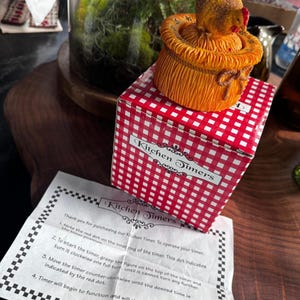 May include: A mechanical kitchen timer shaped like a chicken sitting on a straw bale, packaged in a red and white checkered box labeled Kitchen Timers, accompanied by an instruction sheet with the same text and a black and white border.