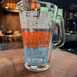 Puede incluir: Una jarra de cristal transparente con un patrón geométrico de rectángulos apilados en bandas horizontales de color blanco, naranja y azul claro. La jarra tiene un asa resistente en el lado derecho y un pico vertedor ancho en el borde superior.